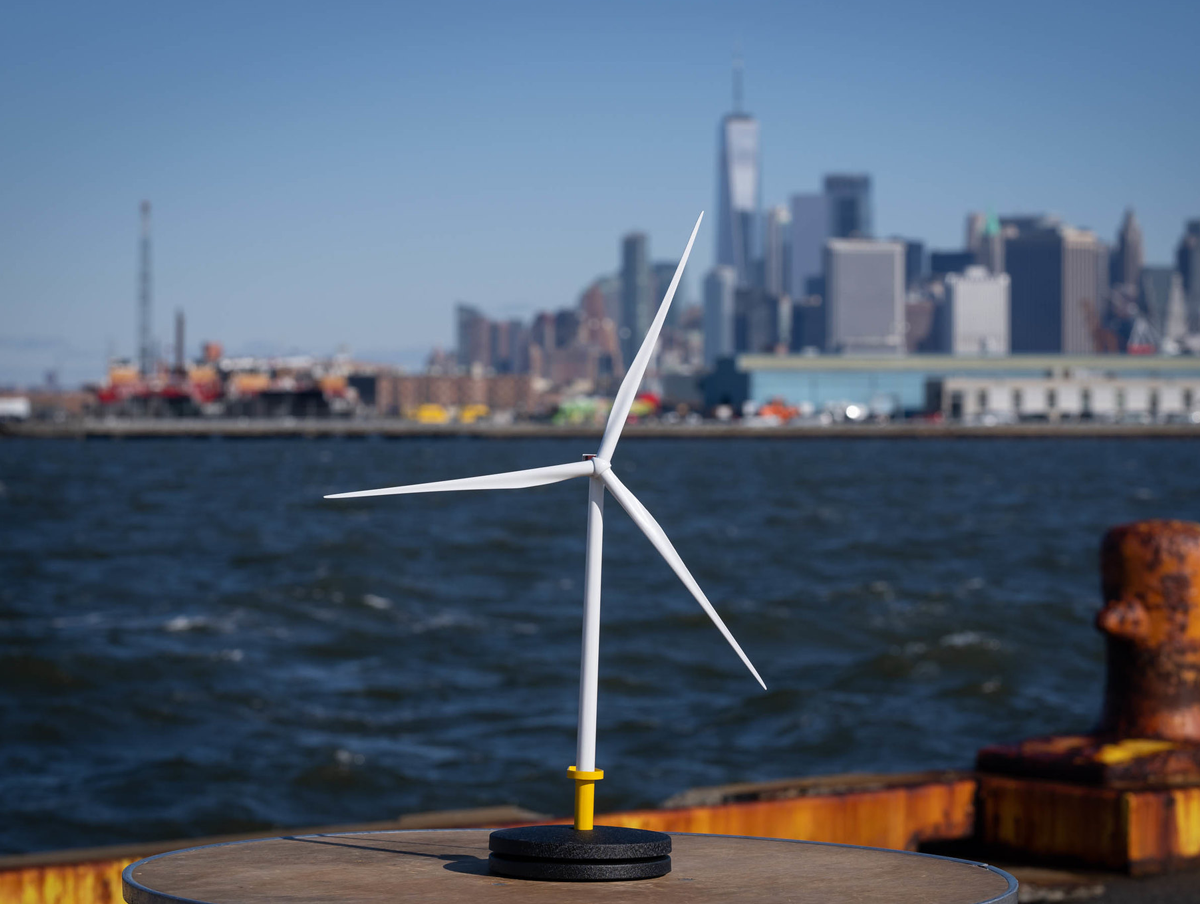 A small wind turbine model is displayed on a table by the water at South Brooklyn Marine Terminal, with a New York City skyline and tall skyscrapers blurred in the background under a clear sky.