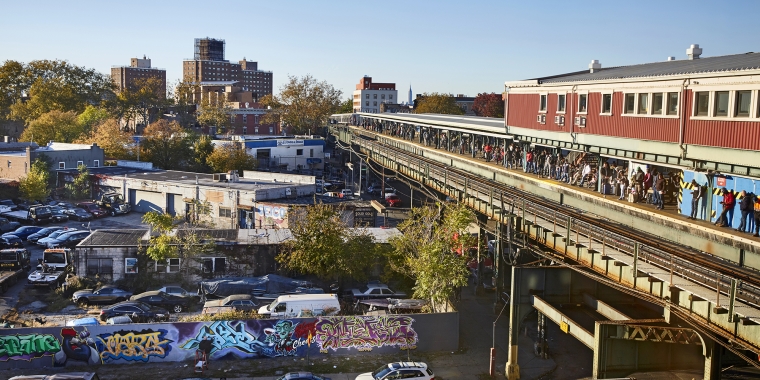 Elevated train platform filled with people runs alongside Broadway Junction, lined with parked cars and colorful graffiti-covered walls. Buildings and trees are visible in the background under a clear blue sky. Photo by John Muggenborg/NYCEDC.