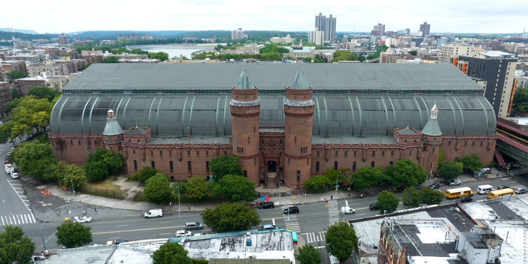 Aerial view of Kingsbridge Armory building with red brick walls and twin turrets, surrounded by NYC streets and greenery. The large, curved metal roof contrasts with the urban landscape, and several vehicles are visible on the roads nearby.