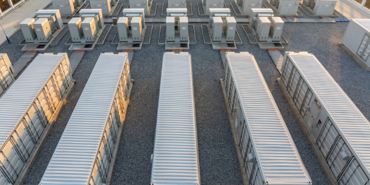 Aerial view of a large battery energy storage power station with rows of white storage containers and electrical equipment on a gravel lot, arranged in an organized grid pattern.