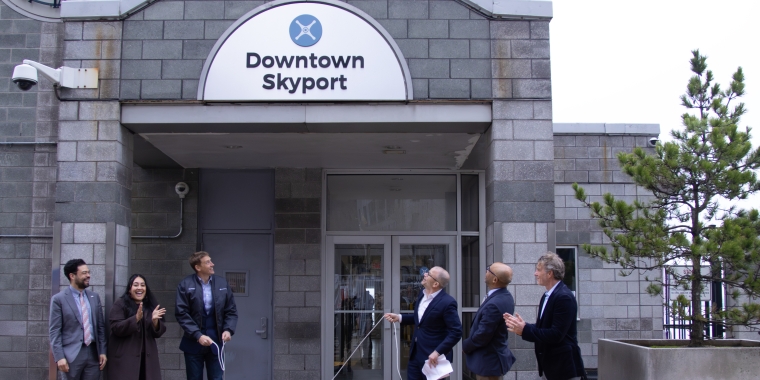 A group of six people, dressed in business attire, stand in front of a building labeled Downtown Skyport. One person is pulling a cord, possibly revealing the sign above the entrance. The setting appears formal and celebratory.